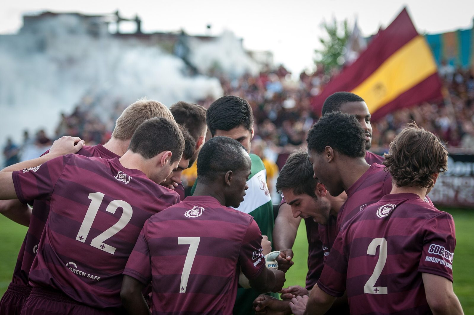 Detroit City players in a huddle with a flag and smoke behind them